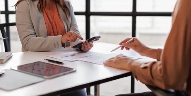 Deux professionnels en réunion autour d'une table, l'une utilise un smartphone, des documents et une tablette devant eux