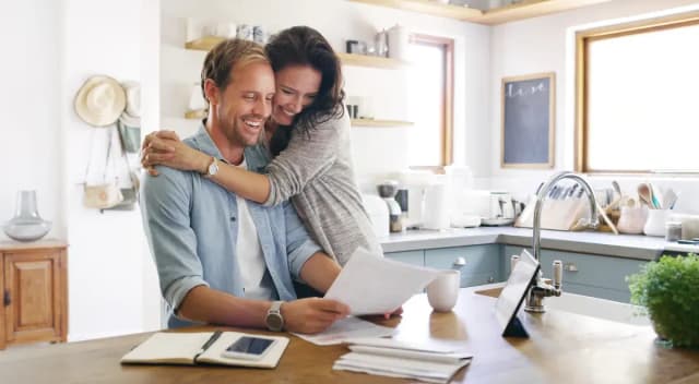 Couple souriant dans une cuisine, la femme enlace l'homme par derrière pendant qu'il lit des documents