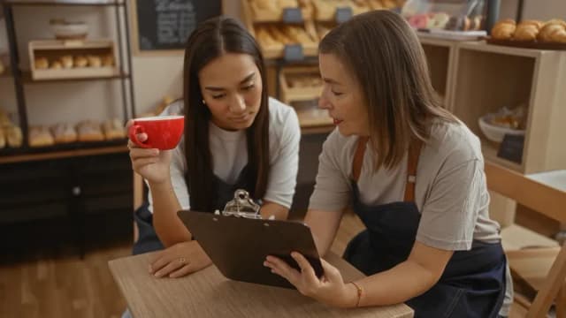 Deux femmes en tablier consultent un document sur un porte-bloc dans une boulangerie, l'une tenant une tasse rouge
