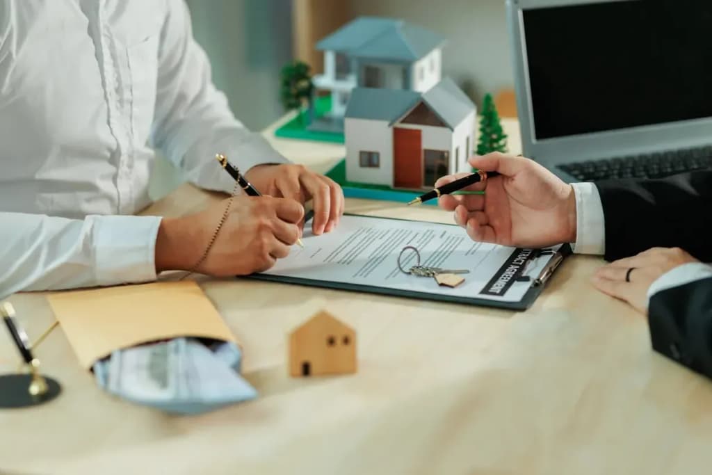 Deux personnes signent un contrat immobilier avec des maquettes de maisons, des clés et un ordinateur