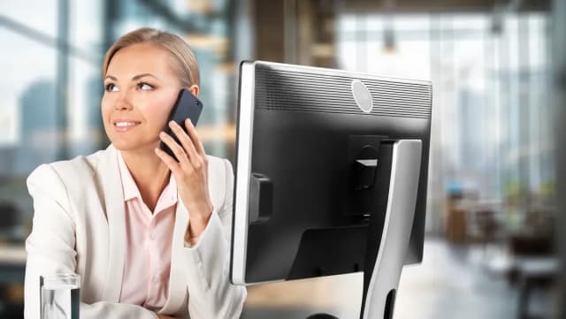Femme en veste blanche souriante téléphonant dans un bureau moderne avec un écran d'ordinateur au premier plan