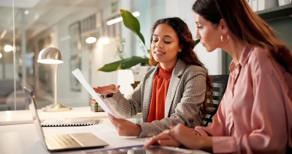 Deux femmes consultent un document dans un bureau moderne, l'une en veste à carreaux l'explique à l'autre