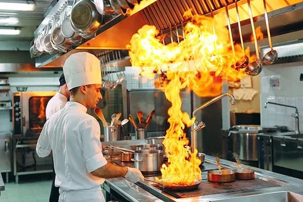 chef en cuisine avec flammes jaillissantes d'une poêle et ustensiles de cuisine en arrière-plan