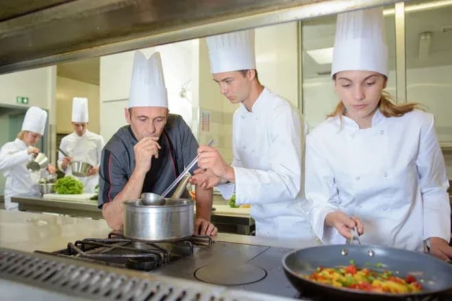 instructeur goûtant une sauce tandis qu'apprentis cuisiniers préparent des plats