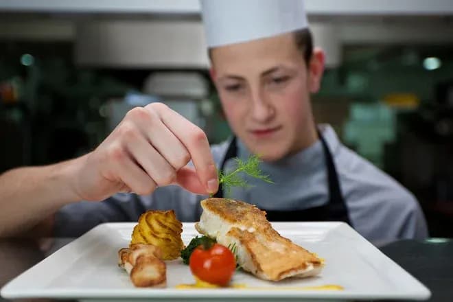 jeune chef ajoute une herbe à un plat de poisson avec garniture sur une assiette blanche