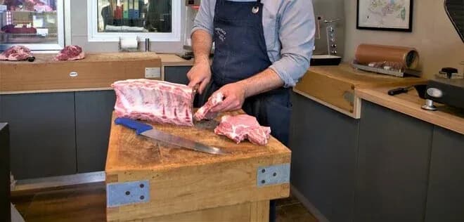 boucher en tablier découpant une côte sur billot en bois dans une boutique