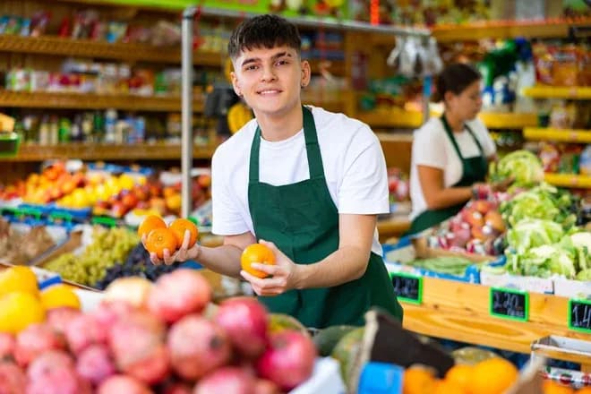 jeune vendeur en tablier vert tenant des oranges devant un étal de fruits