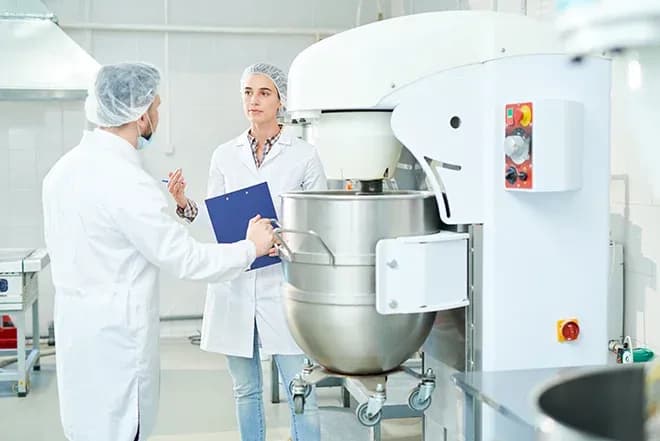 deux personnes en blouses et charlottes discutent près d'un malaxeur industriel