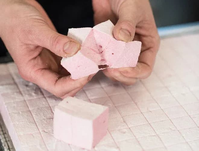 mains tenant des morceaux de guimauve rose sur une surface saupoudrée de sucre glace