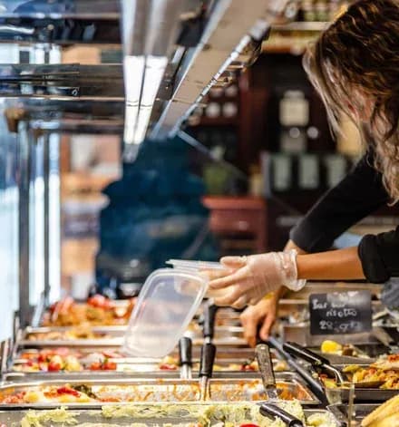 personne gantée remplissant une barquette de plats préparés au comptoir traiteur
