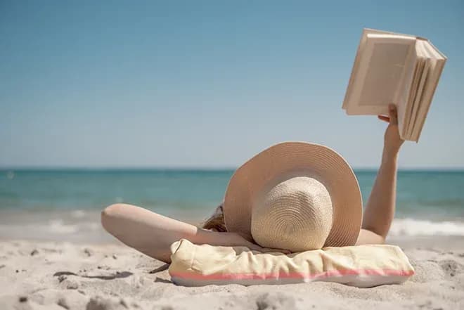 femme allongée sur la plage lisant un livre chapeau de paille et coussin