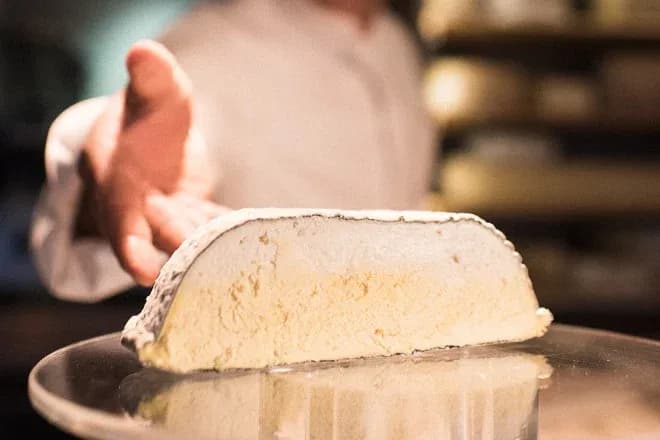 morceau de fromage à croûte fleurie sur plateau en verre, main d'un fromager en arrière-plan