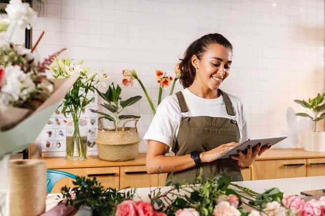 fleuriste souriante utilisant une tablette dans un atelier avec des fleurs autour d'elle