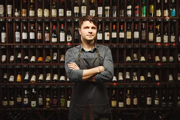 Homme en tablier devant une étagère remplie de bouteilles de vin