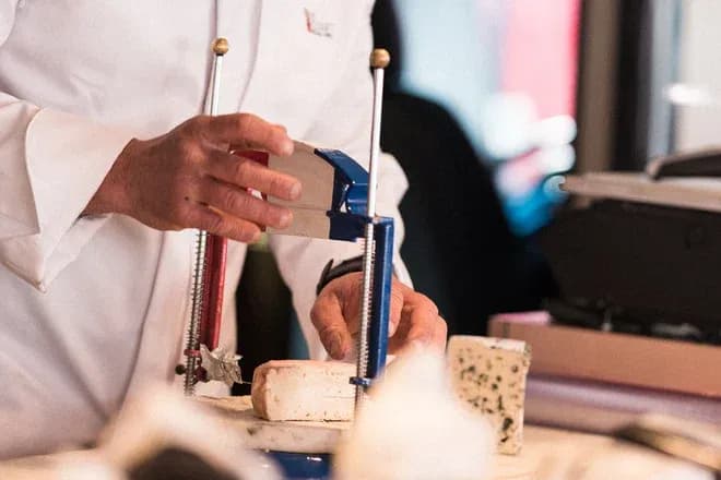 personne en blouse blanche tranchant un fromage avec une lyre professionnelle
