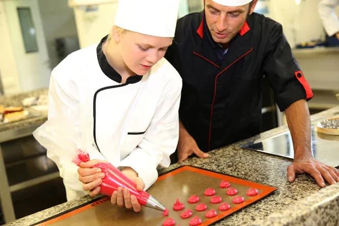 apprentie pâtissière déposant des coques de macarons roses sous la supervision d'un chef