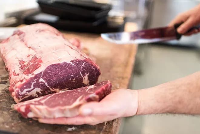 viande rouge sur une planche avec une main tenant une tranche et un couteau à proximité