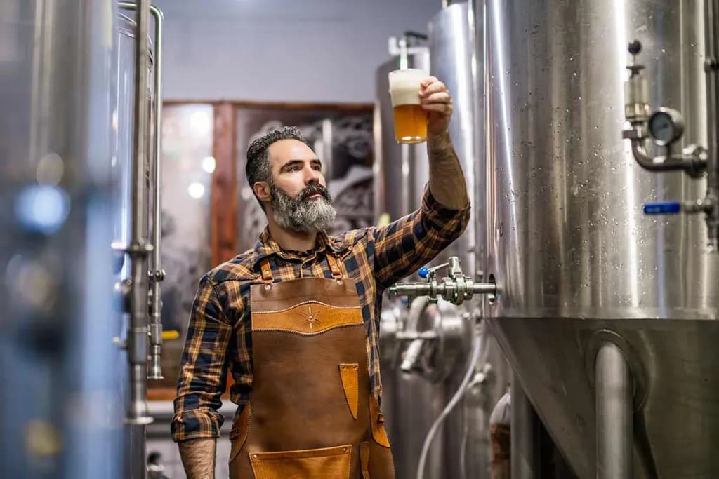 brasseur à la barbe vérifiant la couleur d'une bière devant des cuves inox