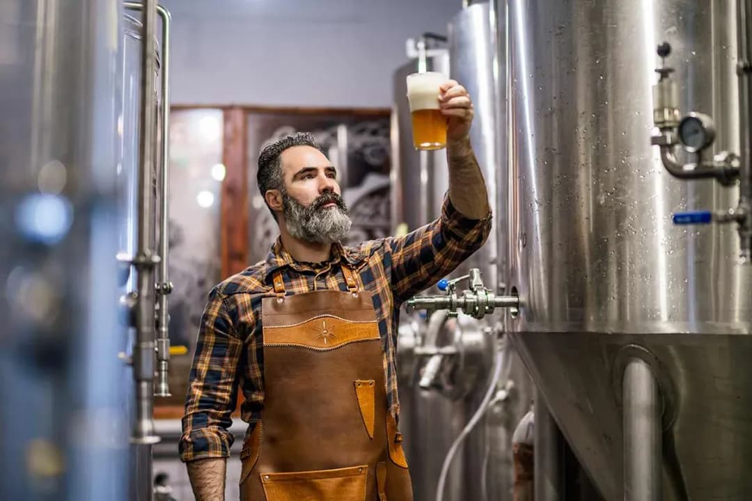brasseur à la barbe vérifiant la couleur d'une bière devant des cuves inox