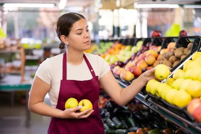 femme en tablier choisissant des pommes dans un étal de fruits frais dans un magasin