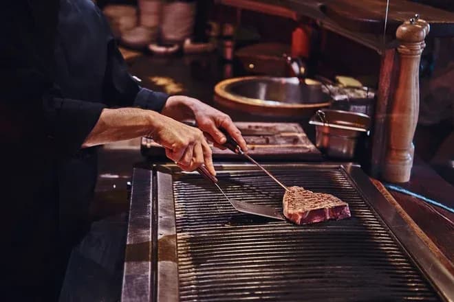 mains d'un chef utilisant spatule et pince pour saisir un steak sur la grille
