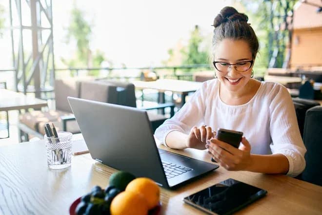 femme souriante consultant son smartphone devant un ordinateur portable au café