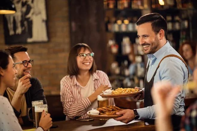 Serveur souriant apportant des plats à des clients dans un bar convivial avec des boissons