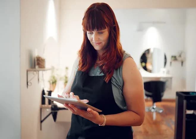 femme avec des cheveux roux utilisant une tablette dans un salon de coiffure moderne