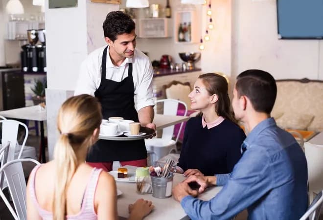serveur sert cafés et pâtisseries à trois clients attablés dans un salon de thé