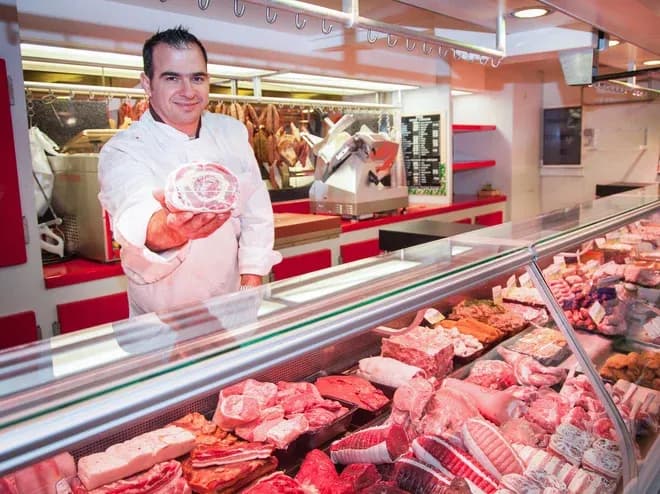 boucher dans un magasin présentant une pièce de viande devant une vitrine remplie de produits carnés