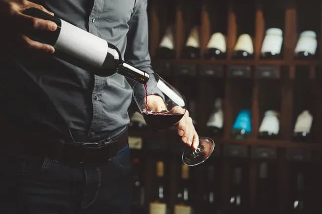 personne versant du vin rouge dans un verre devant une cave à bouteilles
