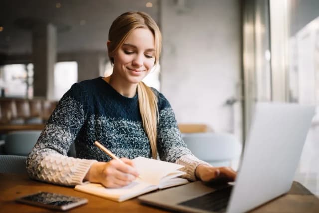 Jeune femme blonde souriante qui prend des notes dans un cahier devant un ordinateur portable dans un café