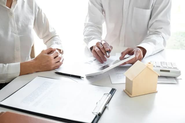 Un conseiller montre des documents à un client, avec une maquette de maison et une calculatrice posées sur un bureau