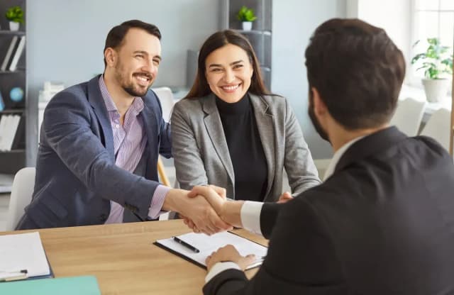 Un homme et une femme souriants serrent la main d'un conseiller en costume, des documents posés sur le bureau