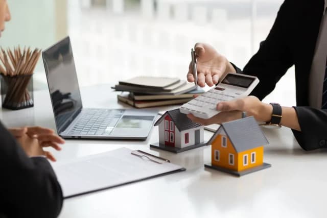 Conseiller tenant une calculatrice face à un client, deux maquettes de maisons et un ordinateur sur le bureau
