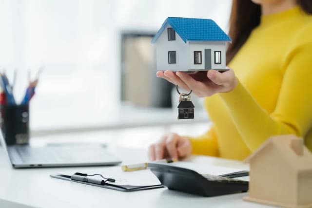 Femme en pull jaune tenant une maquette de maison avec des clés, assise à un bureau avec calculatrice et documents