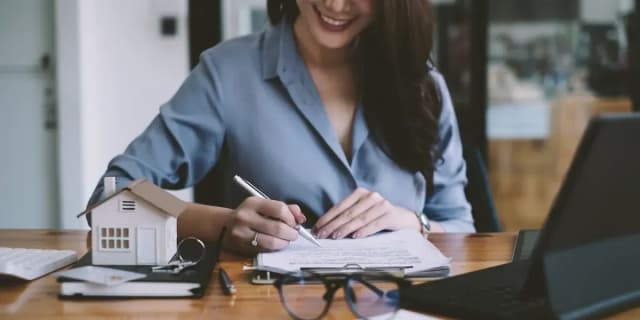 Femme souriante signant des documents immobiliers à un bureau en bois avec une maquette de maison et des clés