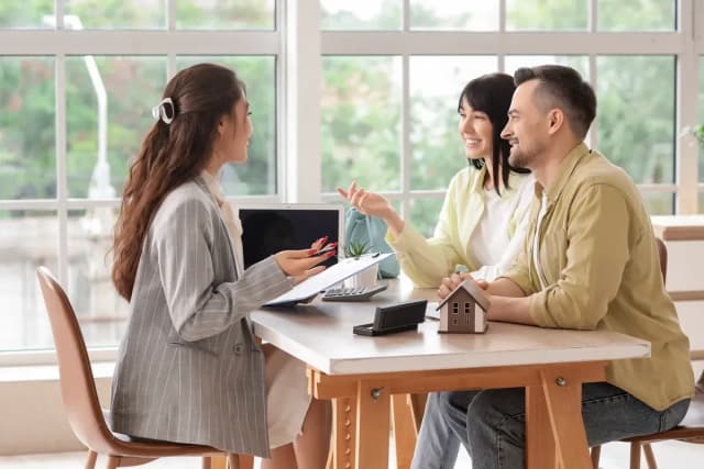 Une conseillère en veste grise explique un dossier à un couple souriant autour d'une table avec une maquette de maison