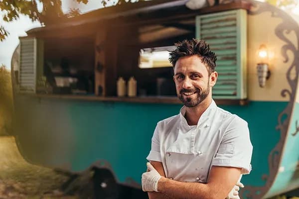 Homme souriant en tenue de chef devant un food truck coloré dans un cadre naturel