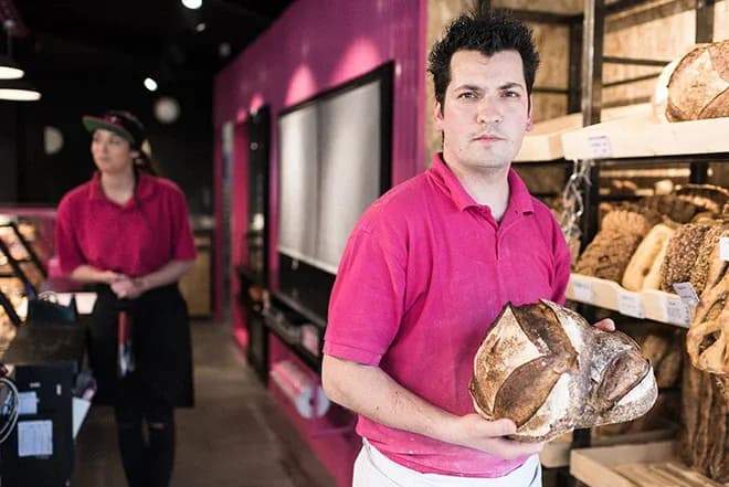 vendeur en polo rose tenant un grand pain rond devant l'étal de la boulangerie