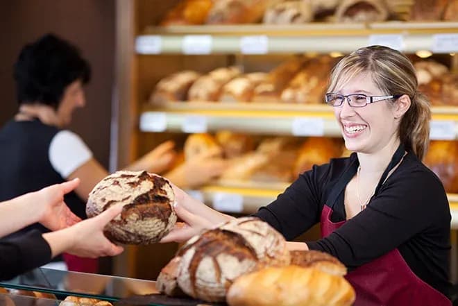 Vendeuse souriante remettant un pain à un client dans une boulangerie bien achalandée