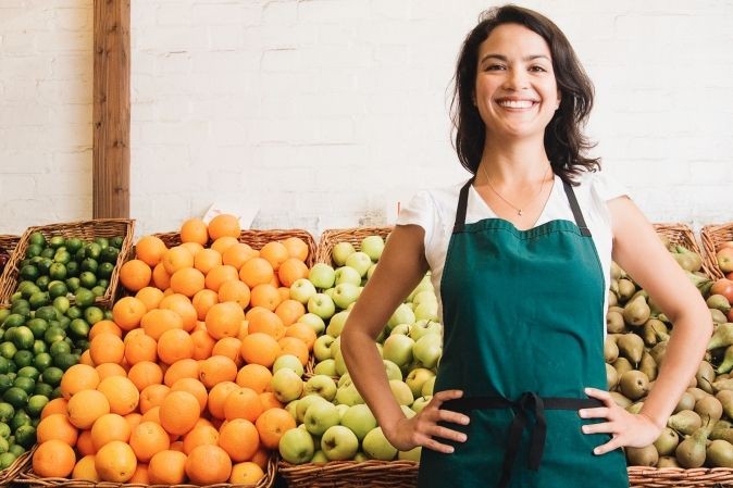 Primeur portant un tablier vert devant son étal de fruits.