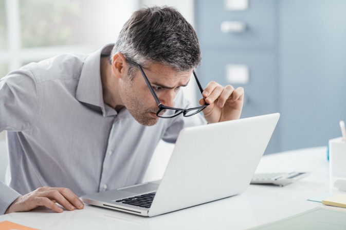 Homme enlevant ses lunettes de vue et se rapprochant de son écran d'ordinateur pour mieux lire